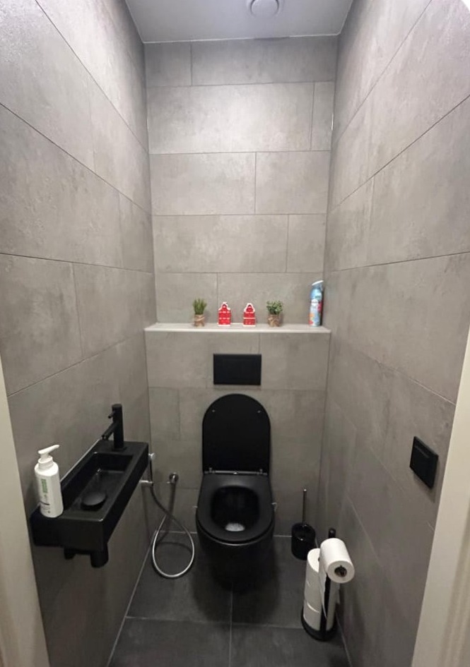 Toilet in grote grijze tegels met een zwart hangend toilet en een zwart fonteintje