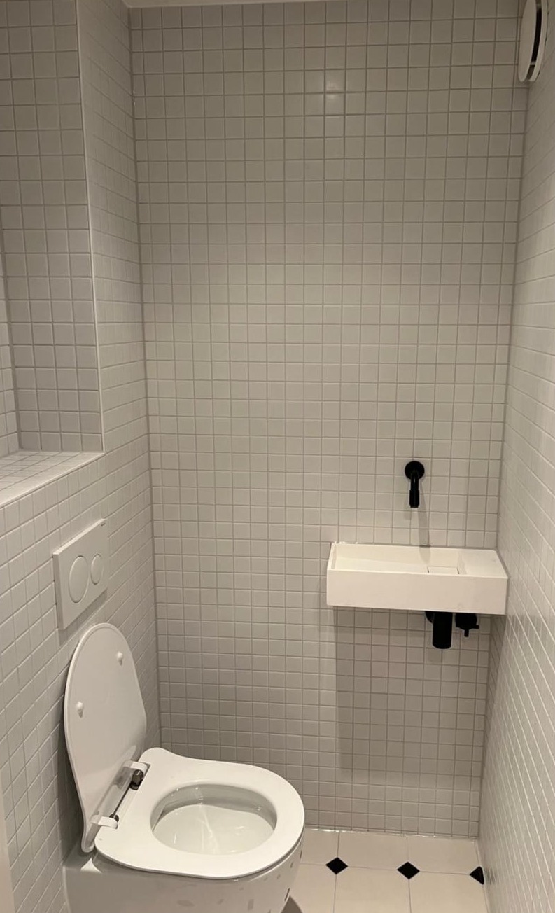 Toilet met witte mozaïektegels, een klein fonteintje en een zwarte kraan